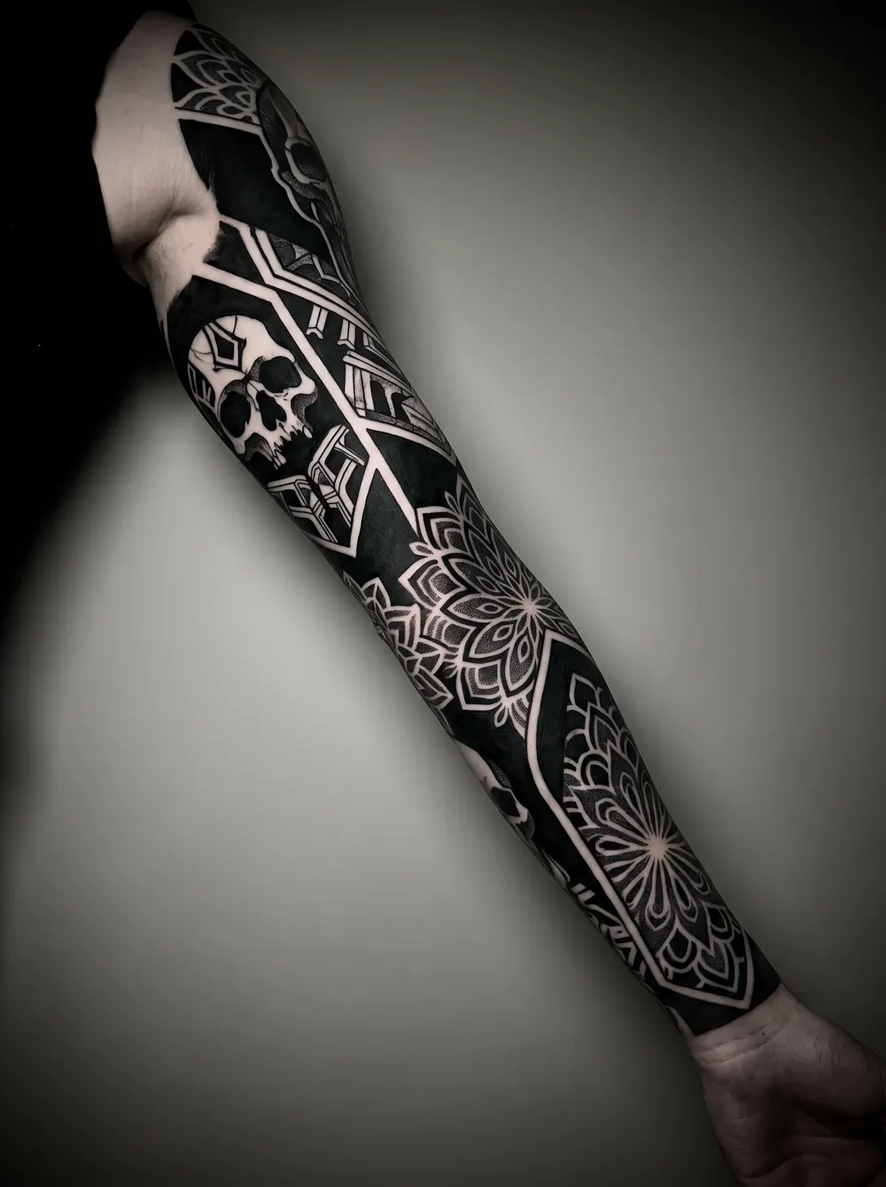A dense black-and-grey blackwork sleeve — solid black fill with negative-space geometry, a skull, and dotwork mandalas — completely burying whatever ink was underneath.