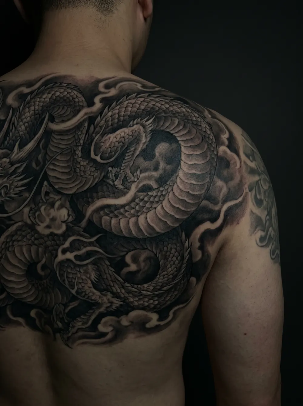 A large black-and-grey realism dragon with layered smoke and clouds sprawling across an upper back and shoulder, dense shadow leaving no light gaps.