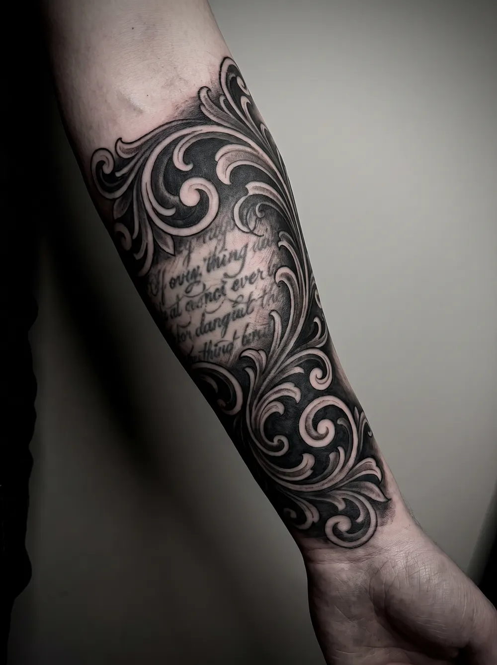 Ornamental black-and-grey scrollwork burying an old script tattoo on a forearm, dark filigree covering the faded lettering.