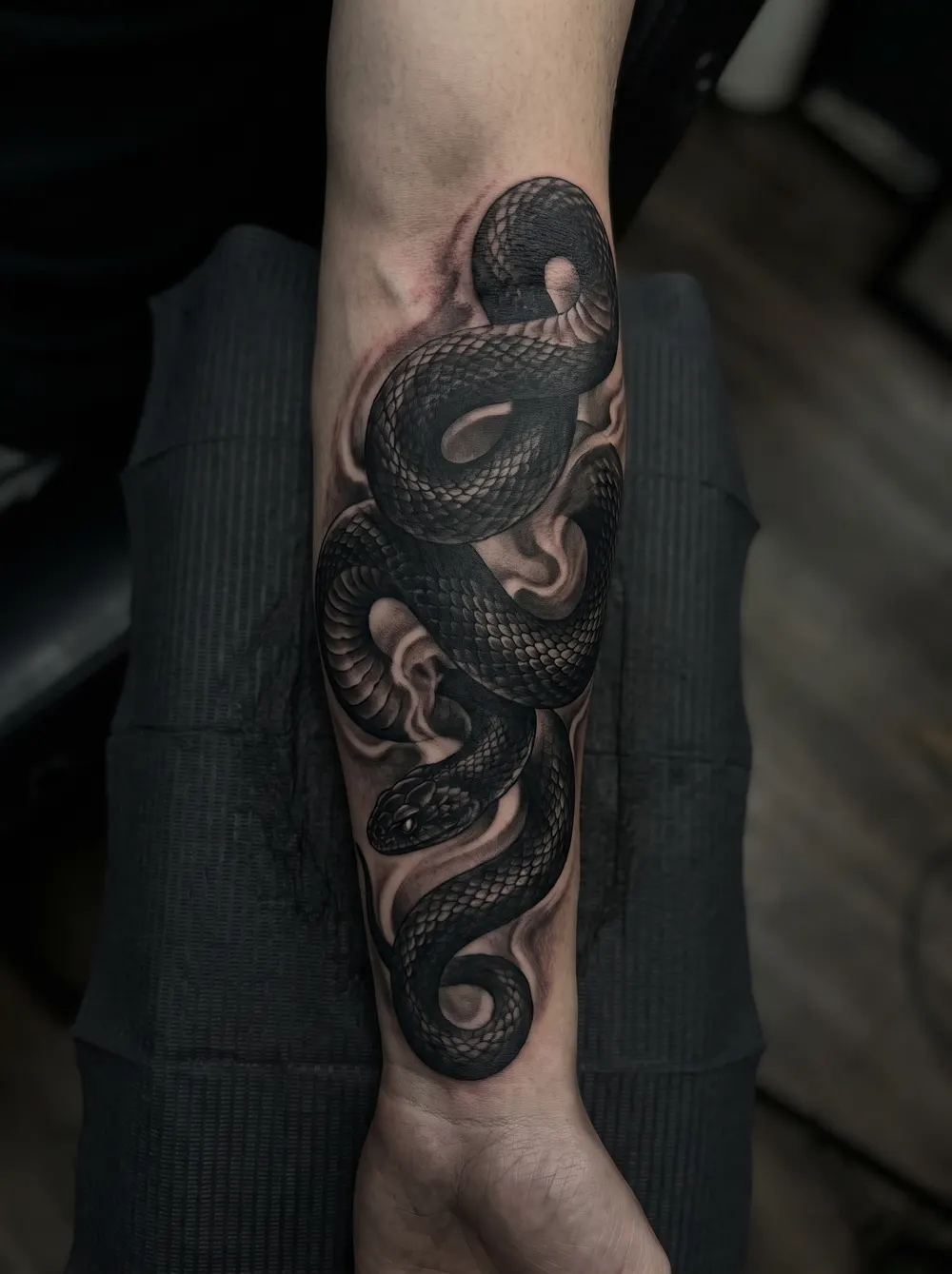 A dense black-and-grey snake cover-up coiling down a forearm, dark scales and shadow engineered to hide older ink.